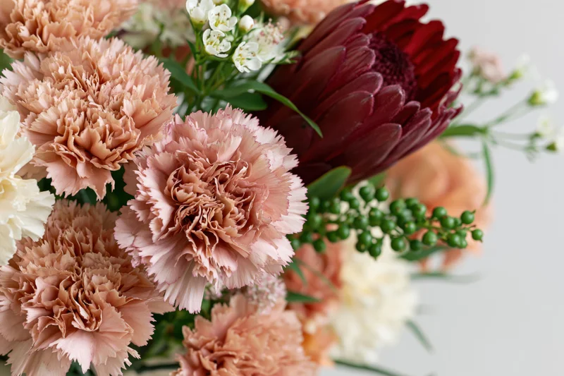 Close-up of a mixed floral arrangement with dusty rose carnations, burgundy dahlia, white filler flowers and greenery on a light background