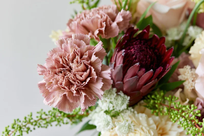 Close-up of a mixed floral arrangement with dusty rose carnations, burgundy artichoke thistle, white filler flowers and green foliage