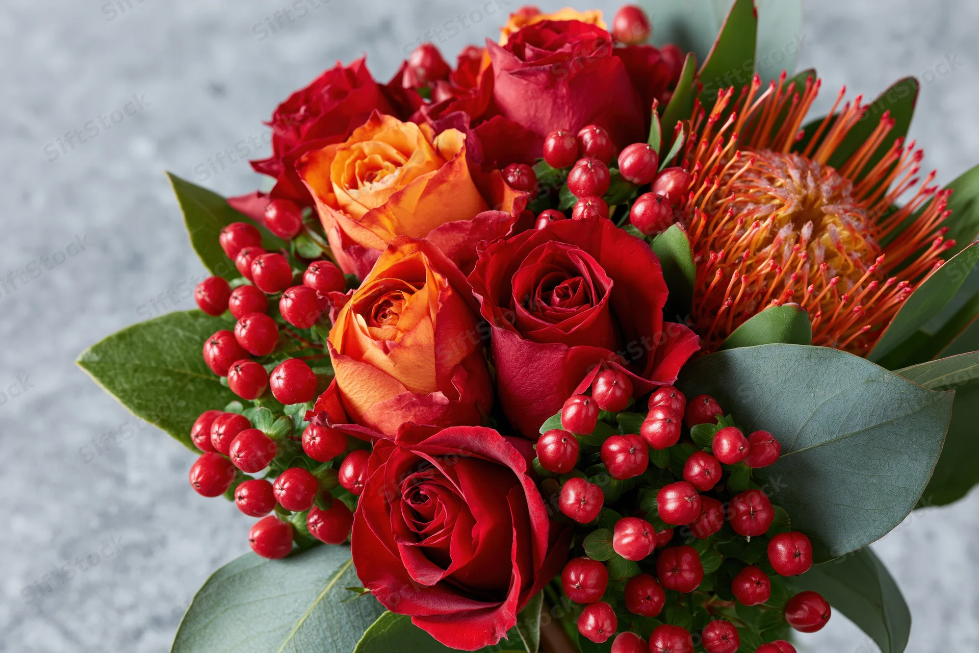 Close-up of a mixed flower bouquet with red and orange roses, protea, berries and green foliage arranged together