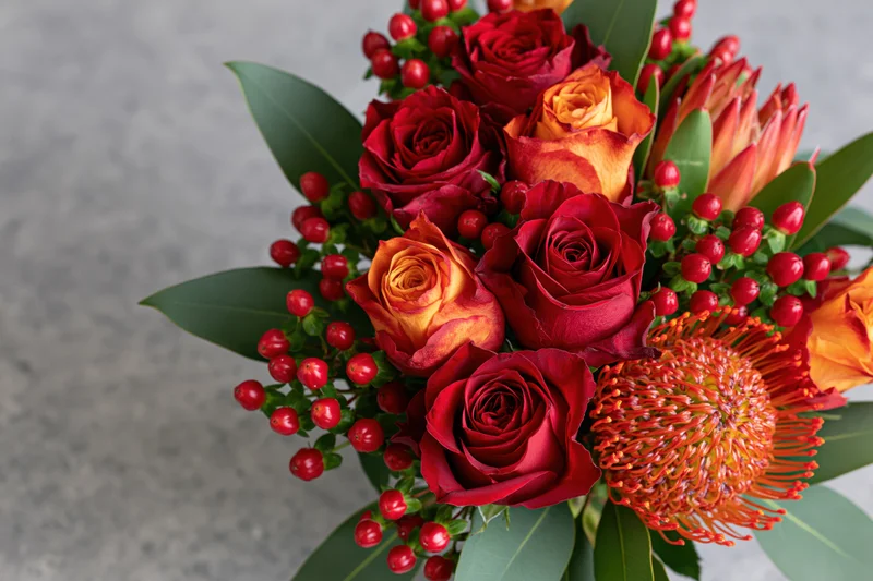 Close-up of a mixed flower bouquet with red and orange roses, protea, berries and green leaves on a gray surface