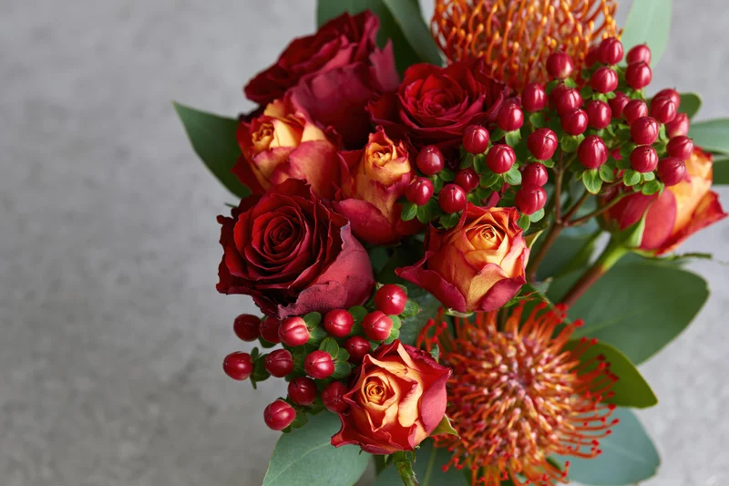 Close-up of a mixed flower bouquet with red and orange roses, pincushion protea, and red berries arranged with green foliage on a neutral background
