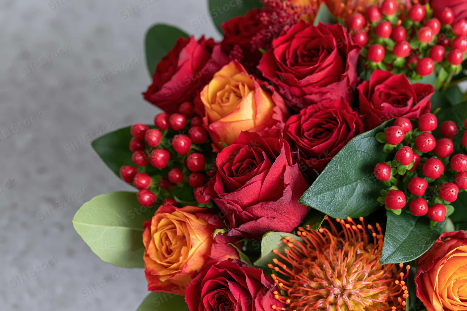Close-up of a mixed flower bouquet with red and orange roses, green leaves, and red berries arranged on a light surface