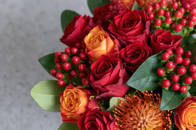 Close-up of a mixed flower bouquet with red and orange roses, green leaves, and red berries arranged on a light surface