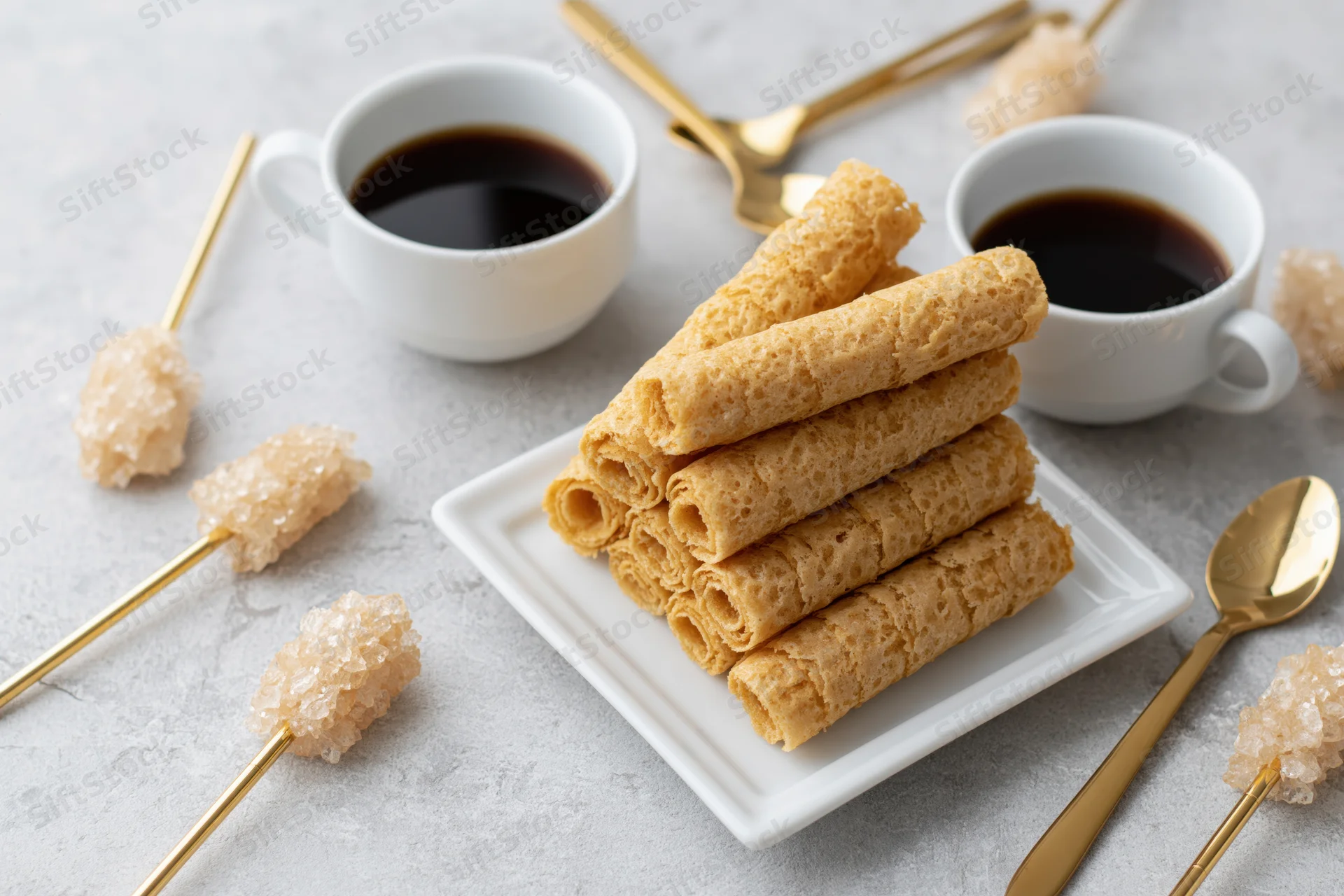 Crispy rolled wafer cookies stacked on a small white square plate with coffee cups and golden spoons nearby
