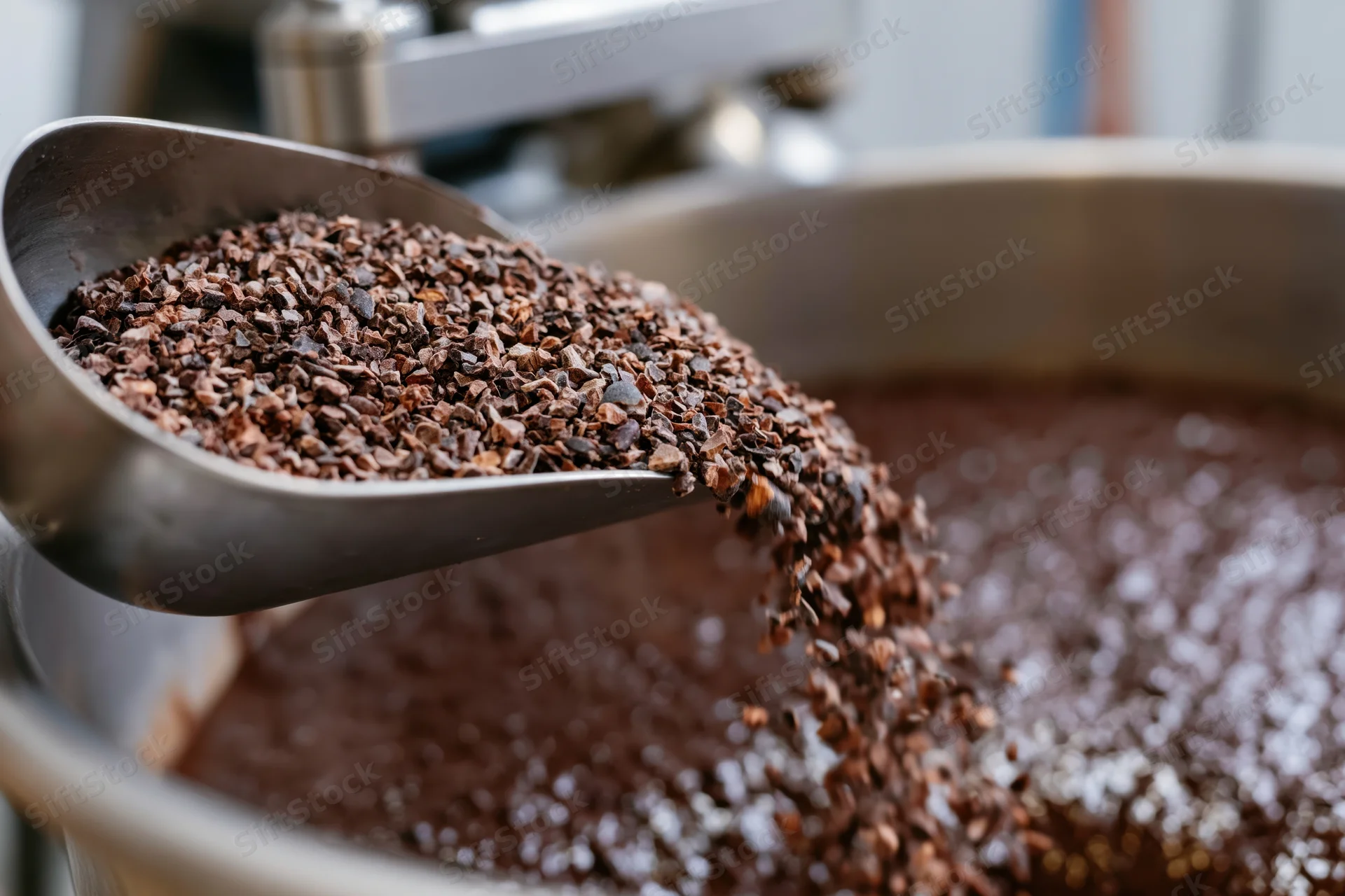 Crushed cacao nibs pouring from a metal scoop into a large industrial mixing vessel