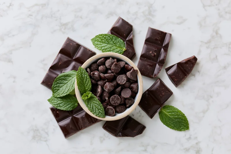Dark chocolate bar pieces broken and scattered around a bowl of chocolate chips with mint leaves on marble surface