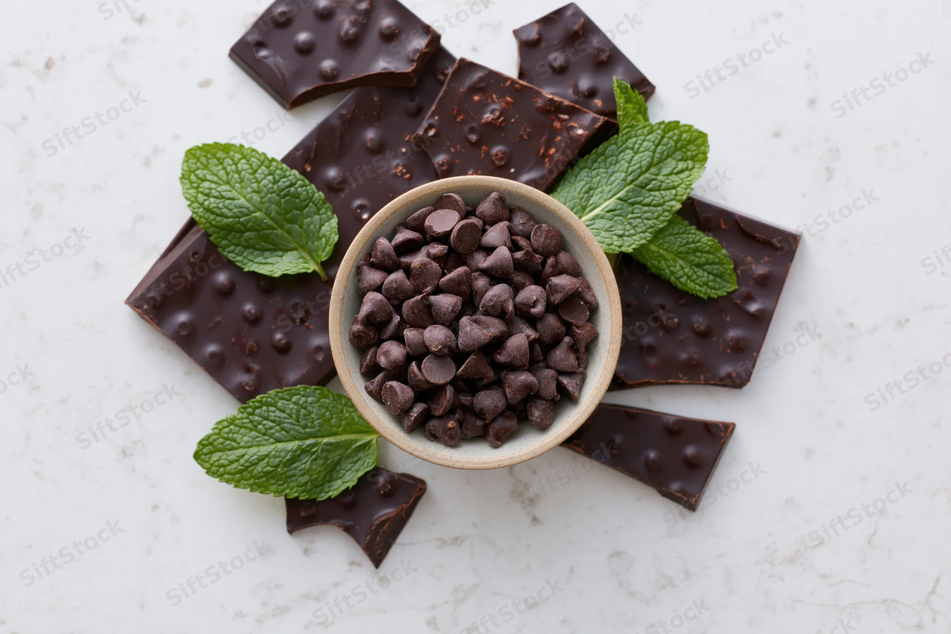 Broken dark chocolate bar pieces scattered around a bowl of chocolate chips with mint leaves on a white marble surface