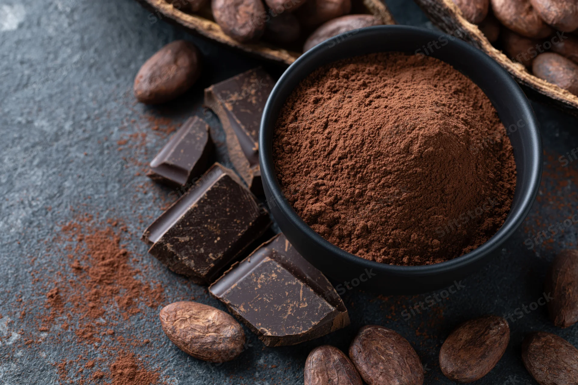 Dark cocoa powder mounded in a matte black bowl surrounded by chocolate chunks and cocoa beans on a textured gray surface