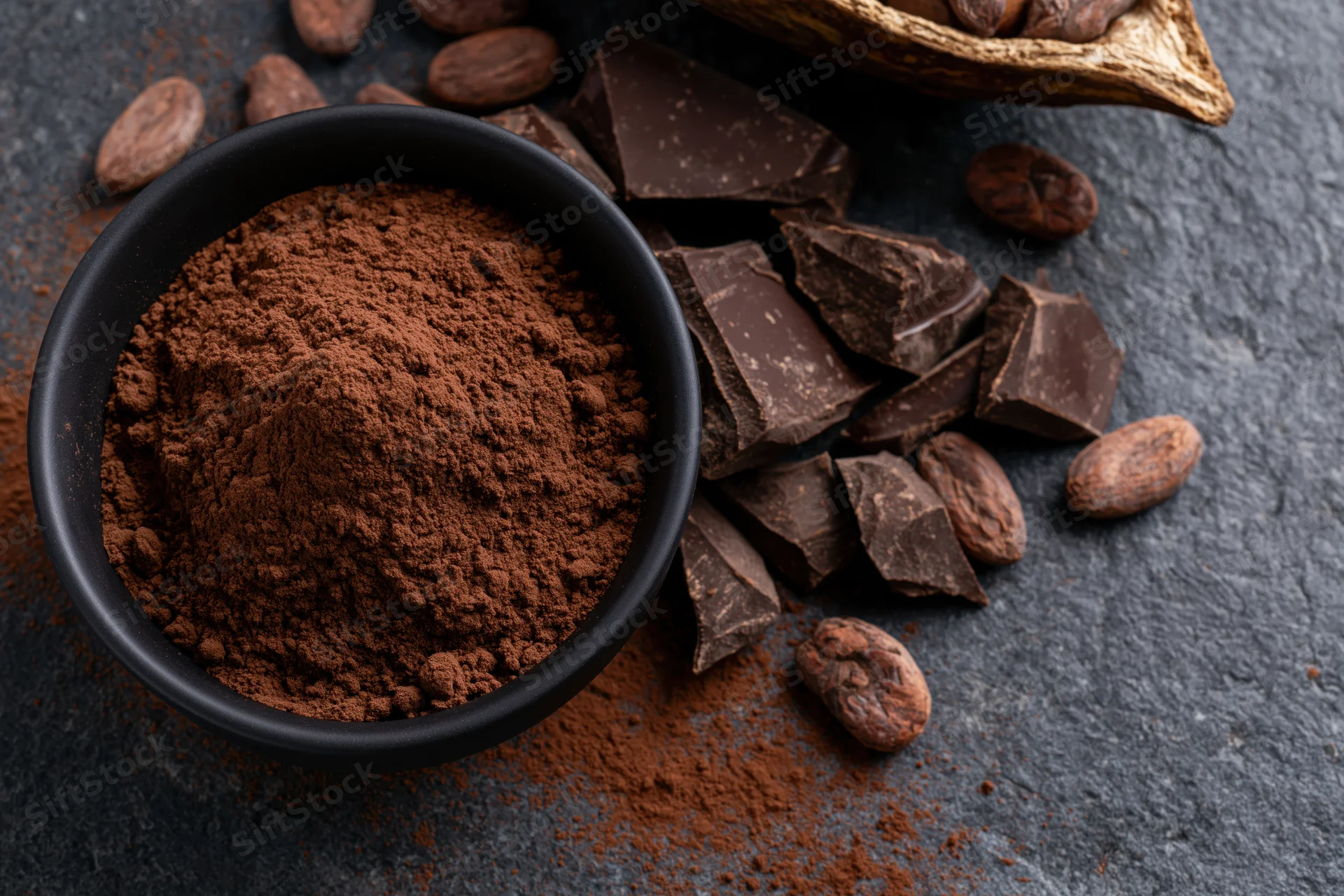 Dark cocoa powder mounded in a matte black bowl surrounded by chocolate chunks and cocoa beans on a slate surface