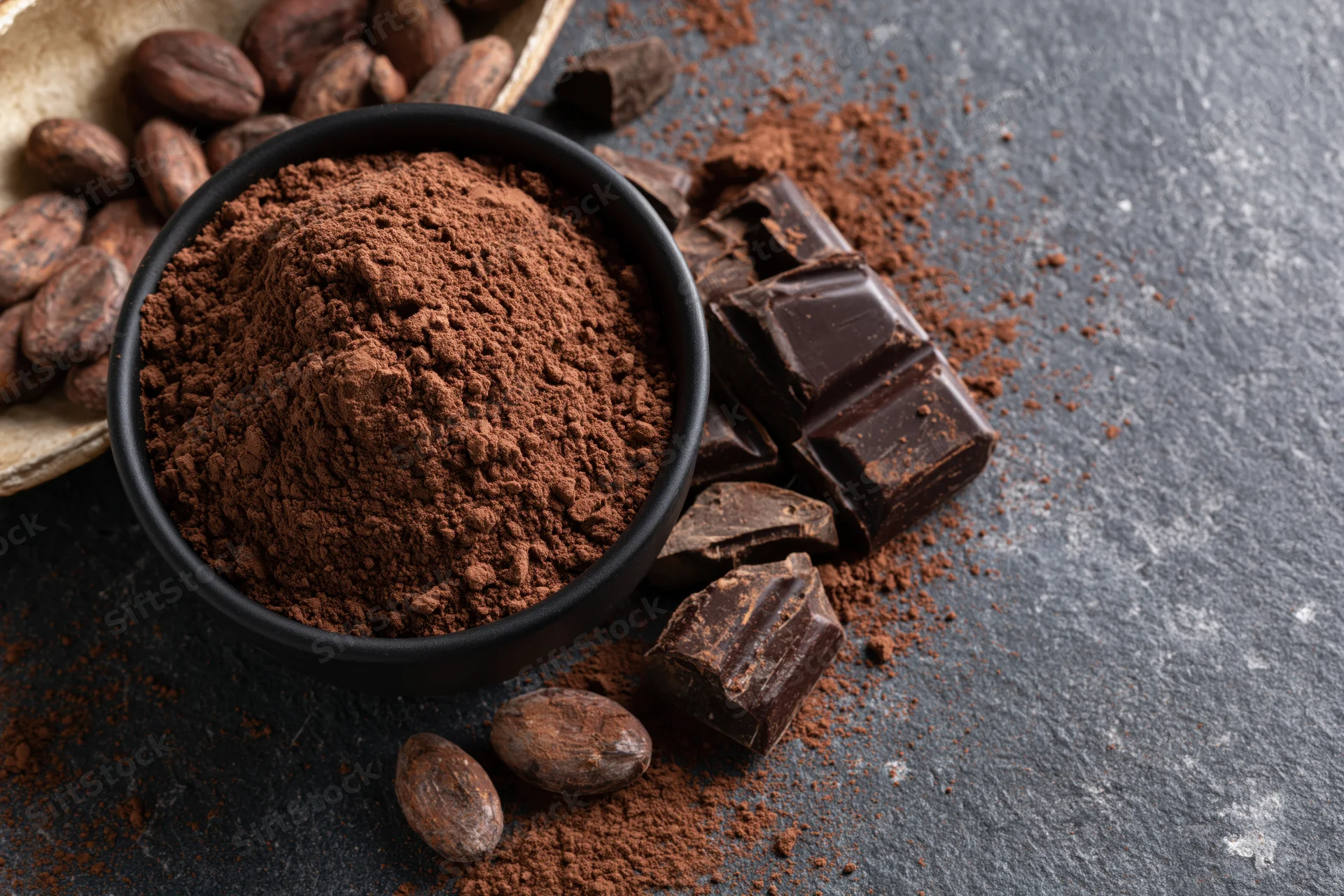 Dark cocoa powder mounded in a matte black bowl surrounded by cocoa beans and broken chocolate pieces on a dark slate surface