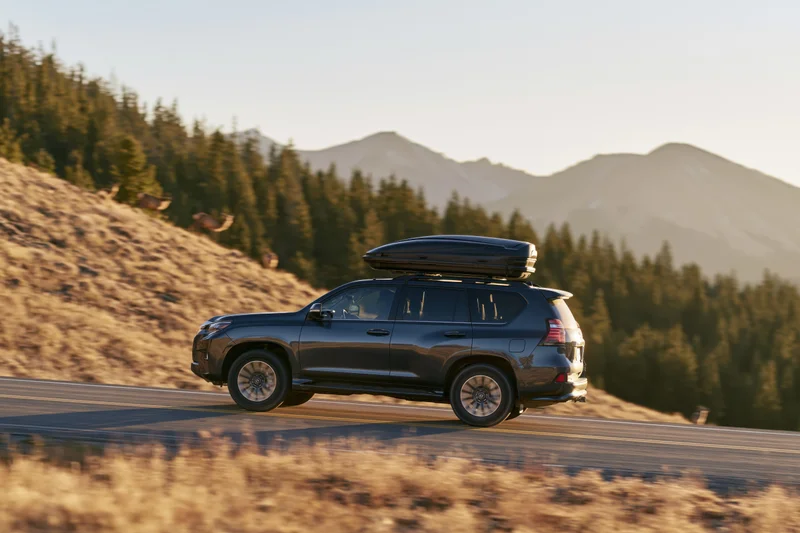 Dark grey luxury suv with black rooftop cargo box driving on winding mountain road surrounded by pine forest and hills at golden hour