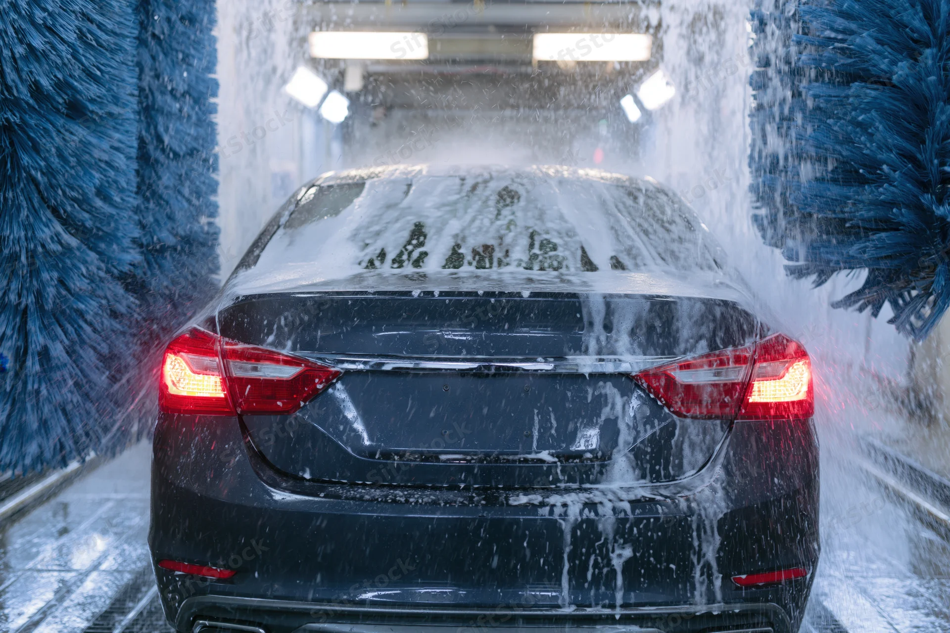 Dark sedan moving through automatic car wash tunnel with foam and blue brushes spraying water
