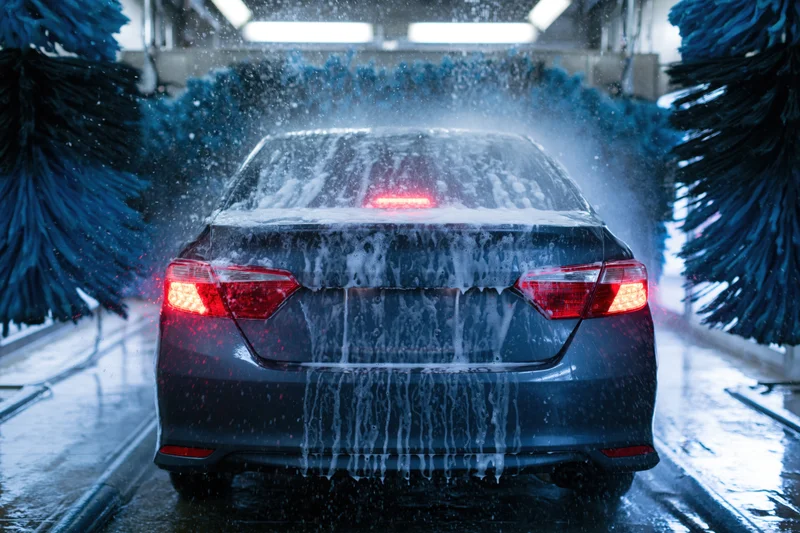 Dark sedan moving through automatic car wash tunnel viewed from rear with foam and water spraying