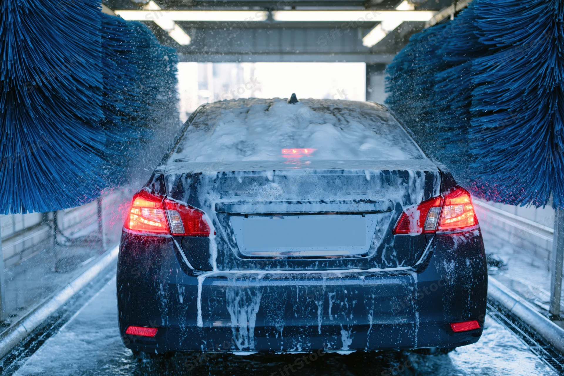 Dark sedan moving through automatic car wash tunnel with rotating blue brushes and soap foam covering rear view