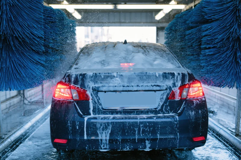 Dark sedan moving through automatic car wash tunnel with rotating blue brushes and soap foam covering rear view