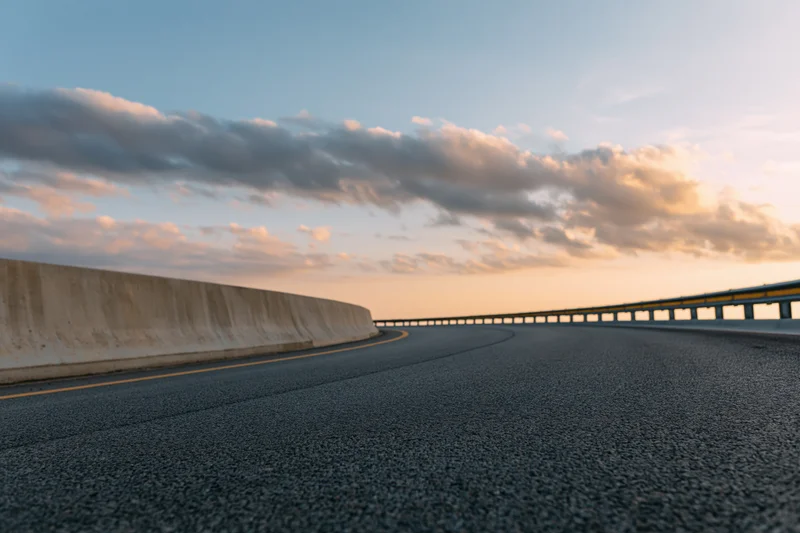 Curved asphalt highway sweeping right with concrete barrier and guardrail under sunset sky