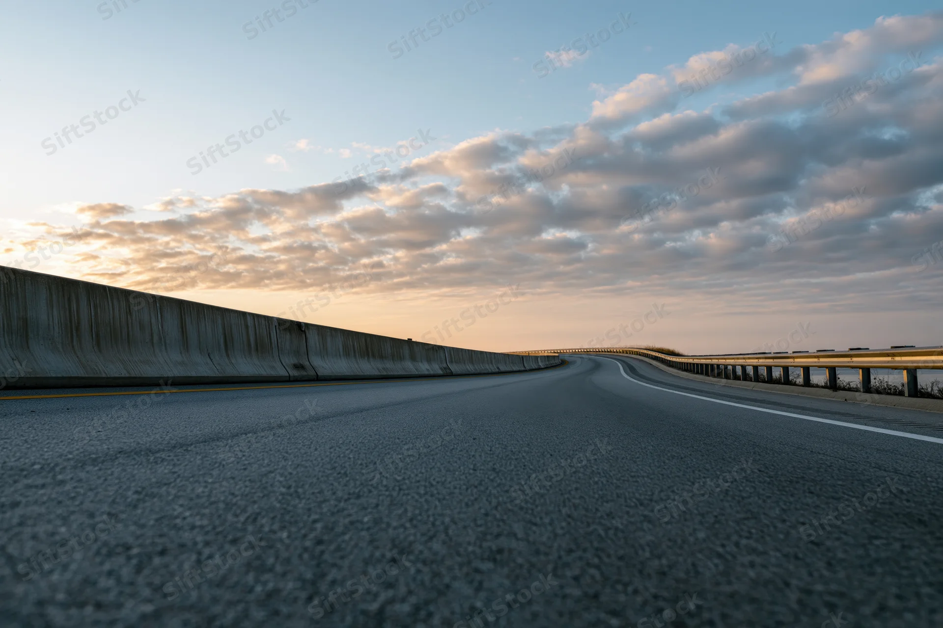 Empty curved highway sweeping to the right with guardrails and cloudy sky at dusk