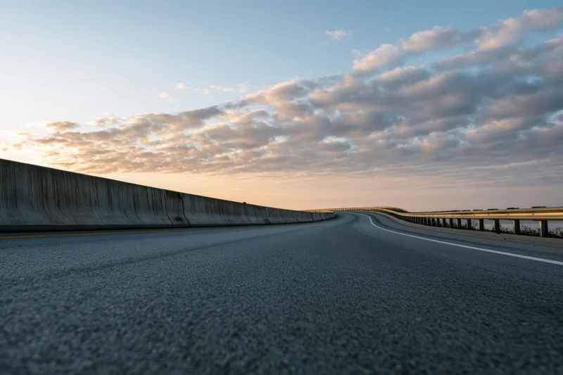 Empty curved highway sweeping to the right with guardrails and cloudy sky at dusk