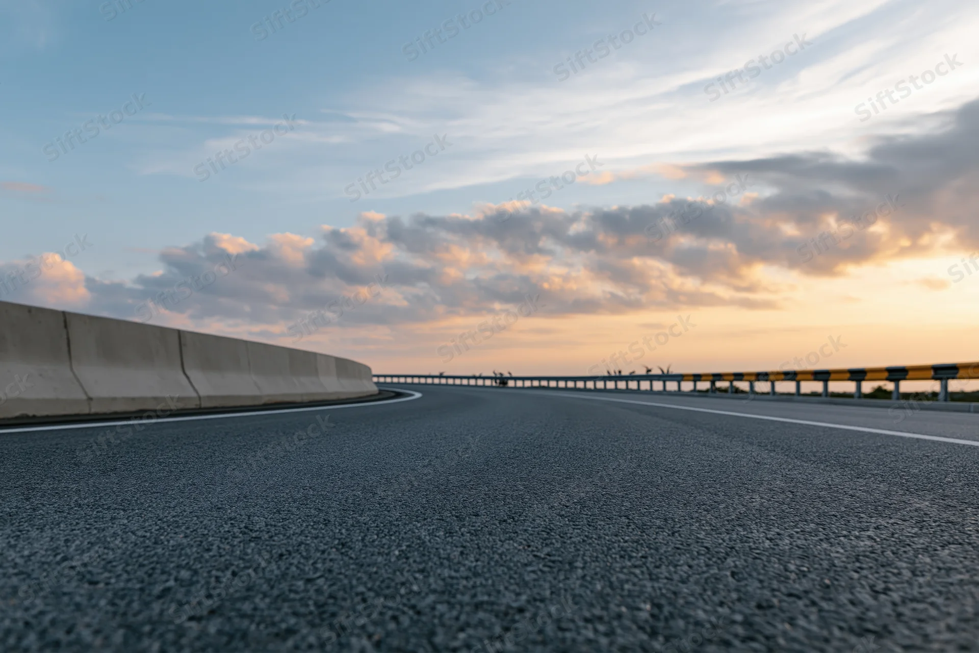 Curved asphalt highway sweeping right with concrete barrier and guardrail under sunset sky