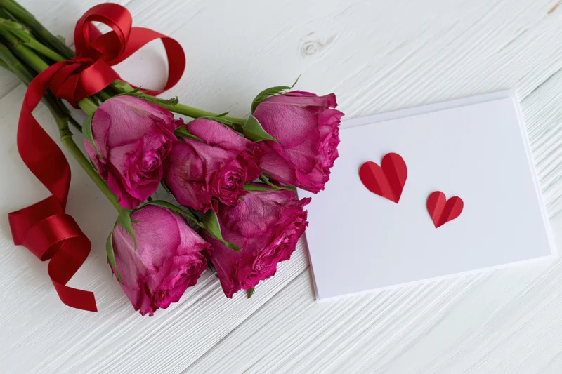 Five deep pink roses tied with a curled red satin ribbon laid on a white wooden surface beside a blank card with two red paper hearts