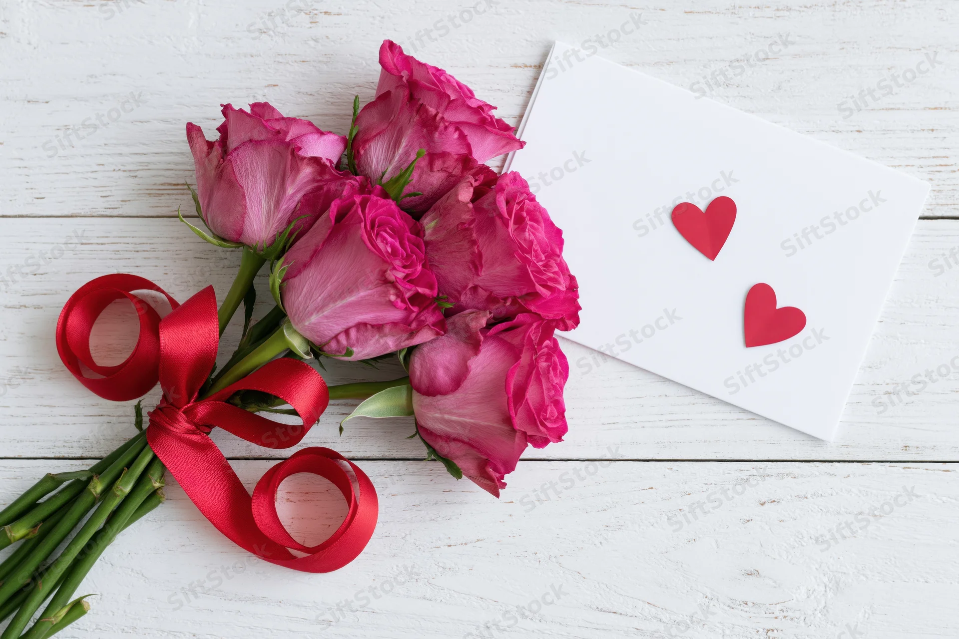Bouquet of pink roses tied with red satin ribbon on white wooden surface beside a white card with red hearts