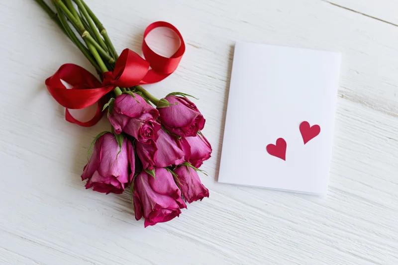 Five deep pink roses tied with a curled red satin ribbon on a white wooden surface beside a white card with two red hearts