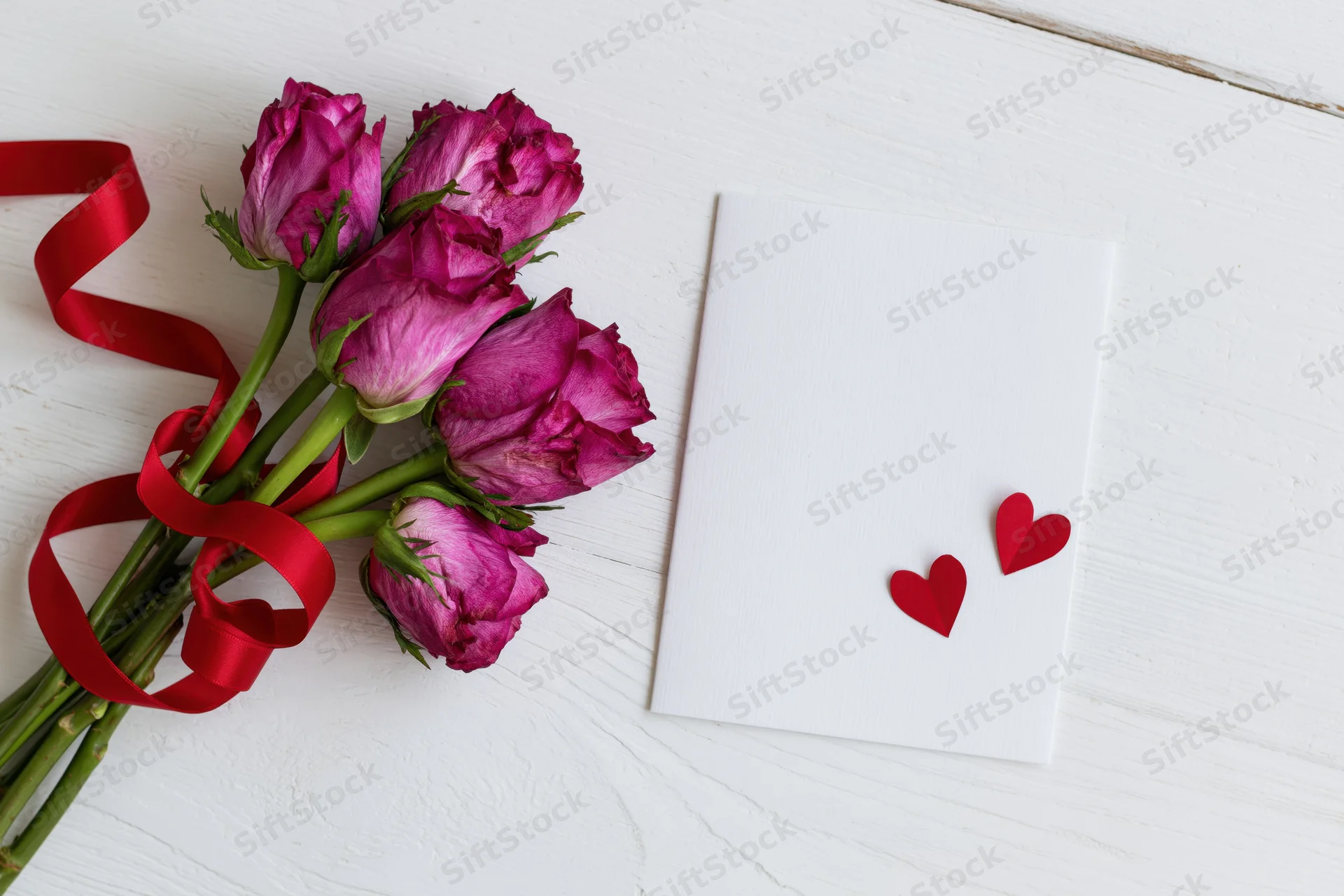 Bouquet of deep pink roses tied with red satin ribbon on white wooden surface beside blank card with two red hearts