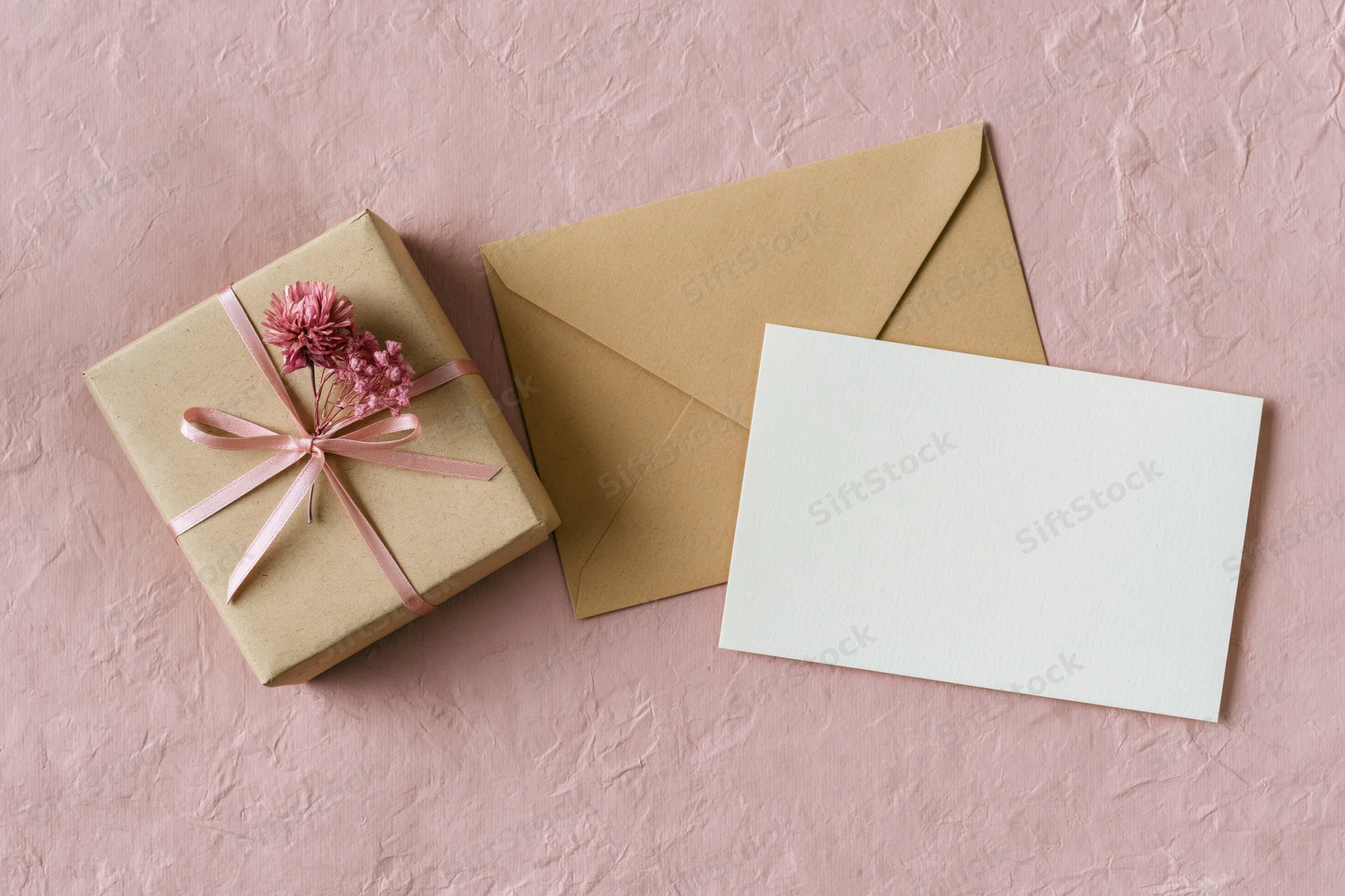 Kraft paper gift box tied with pink ribbon and dried flowers beside envelope and blank card on pink textured surface