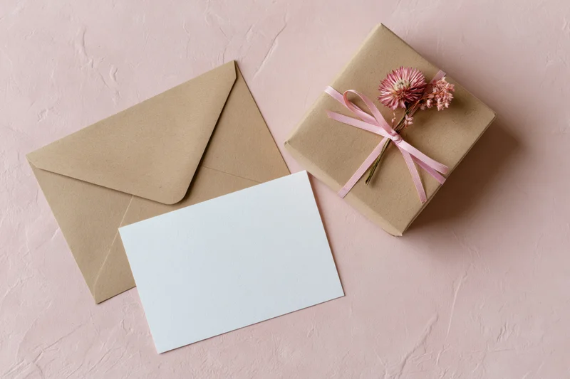 Kraft paper gift box tied with pink ribbon and dried flower beside envelope and blank card on pink surface