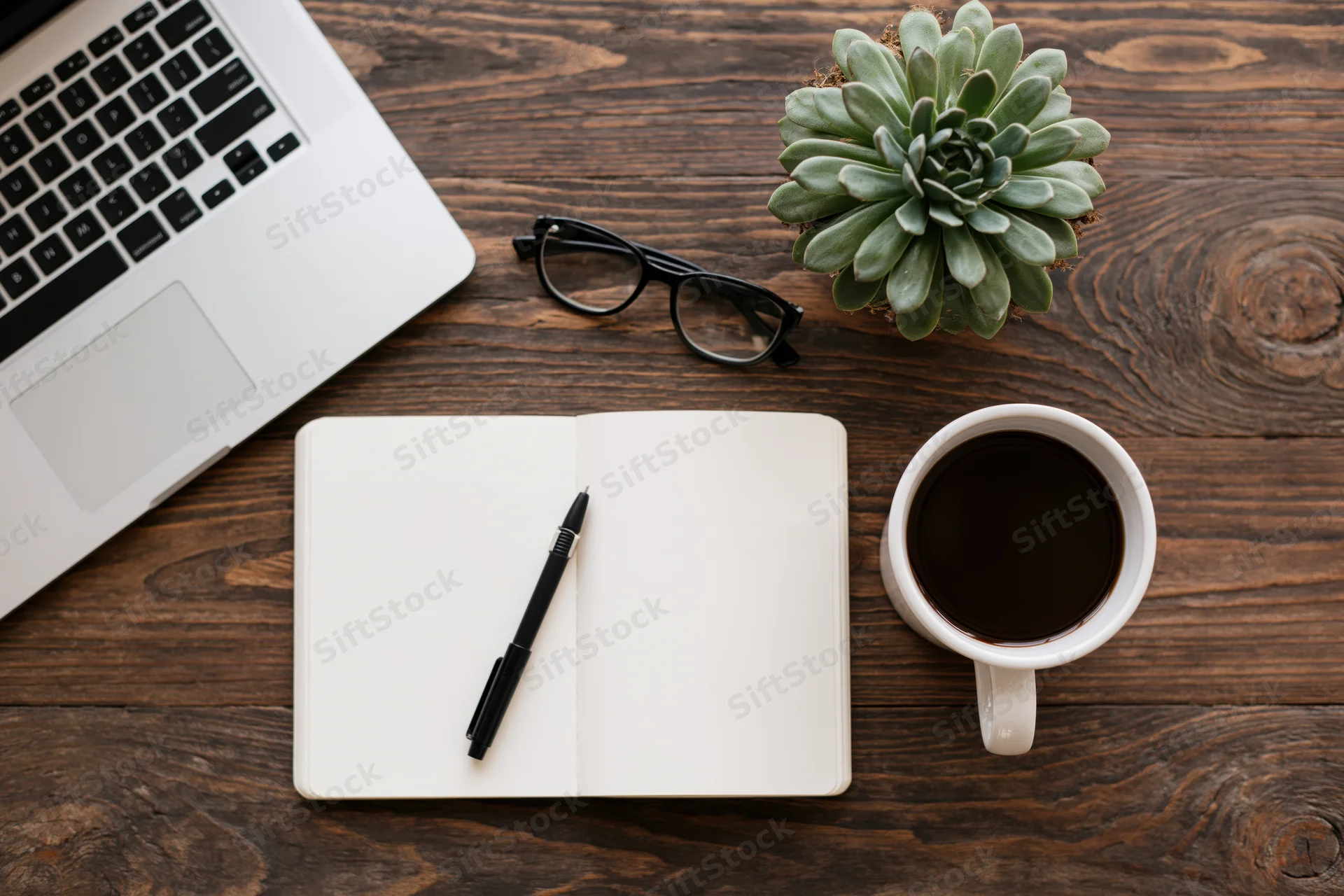 Overhead view of a wooden desk with a silver laptop, open notebook, pen, coffee mug, glasses, and succulent plant arranged for work or study