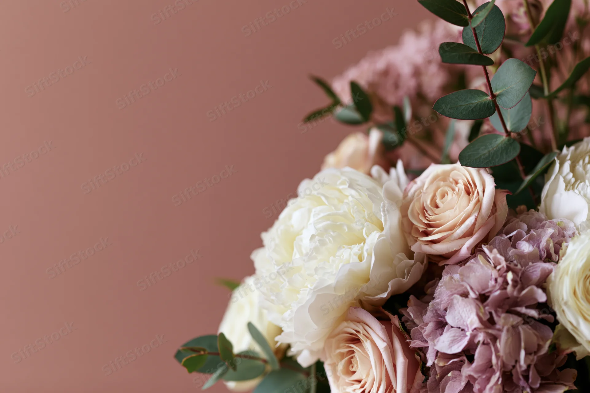 Floral arrangement of white peonies, blush roses, and lavender hydrangeas with eucalyptus foliage against a muted pink background