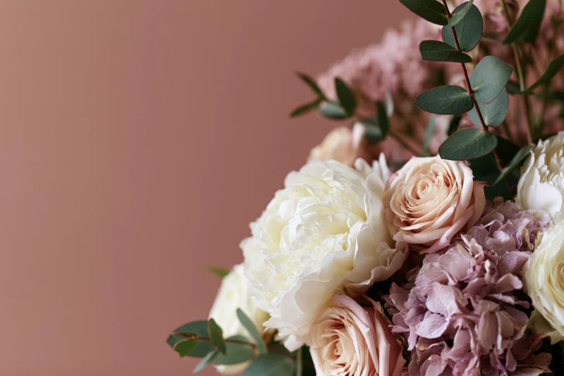 Floral arrangement of white peonies, blush roses, and lavender hydrangeas with eucalyptus foliage against a muted pink background
