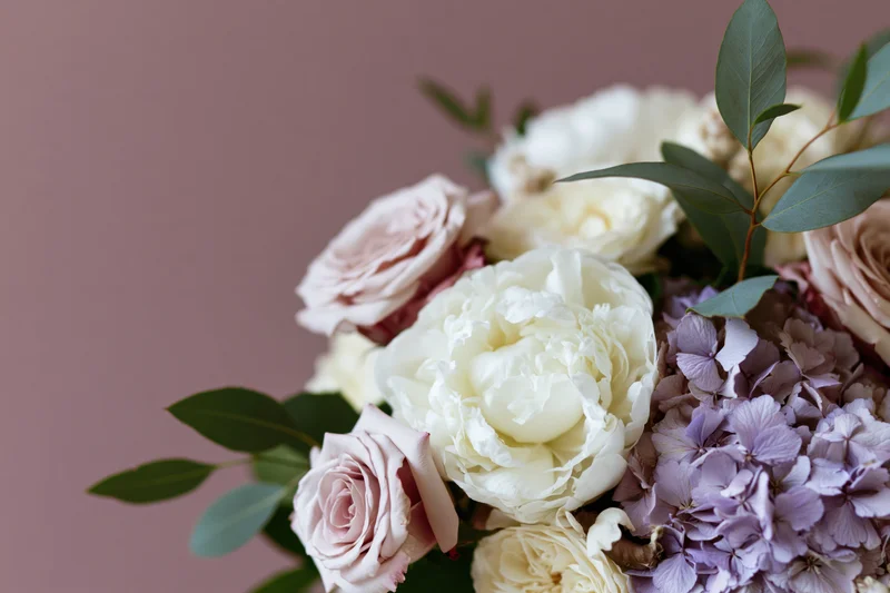 Floral arrangement of white peonies, blush roses, and lavender hydrangeas with green foliage against a muted pink background