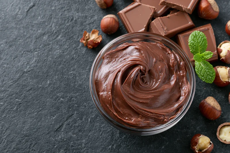Swirled chocolate hazelnut spread in a glass bowl surrounded by chocolate pieces and hazelnuts on a dark stone surface