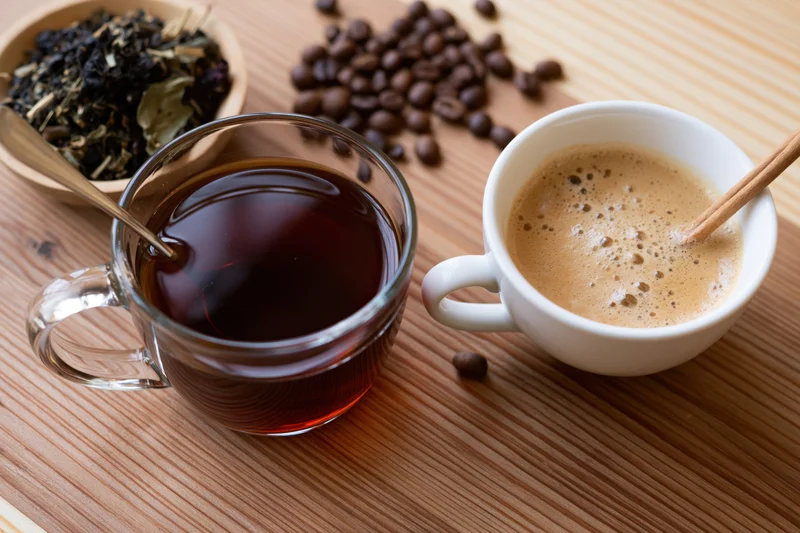 Glass mug of dark tea and white cup of frothy coffee on wooden table with coffee beans and tea leaves nearby