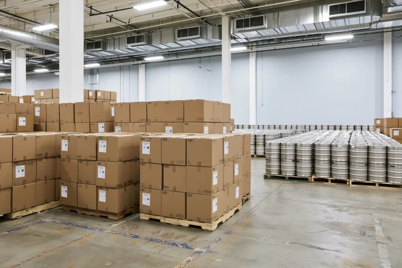Tall stacks of cardboard boxes and metal kegs arranged on pallets in a large warehouse interior
