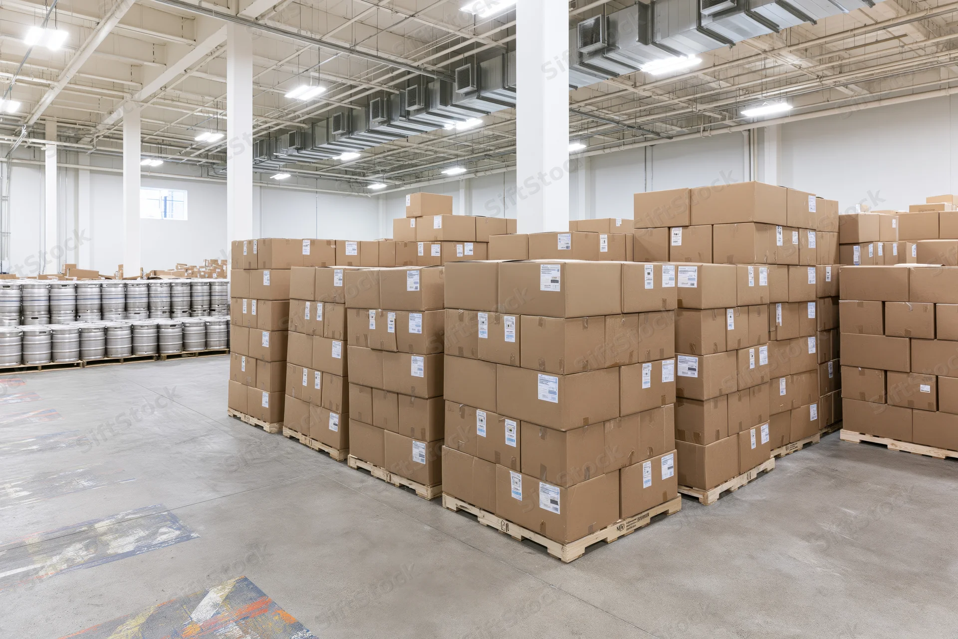 Tall stacks of cardboard boxes on pallets arranged in rows inside a large commercial warehouse with metal barrels in the background
