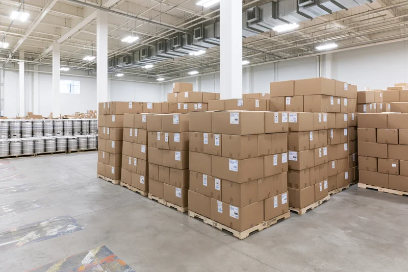 Tall stacks of cardboard boxes on pallets arranged in rows inside a large commercial warehouse with metal barrels in the background