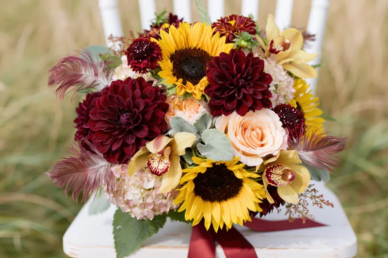 Lush autumn bridal bouquet with sunflowers, dahlias, and feathers resting on a white wooden chair outdoors