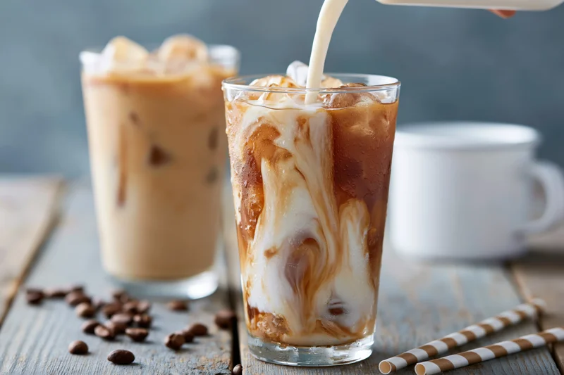Milk being poured into a tall glass of iced coffee with ice cubes on a wooden table
