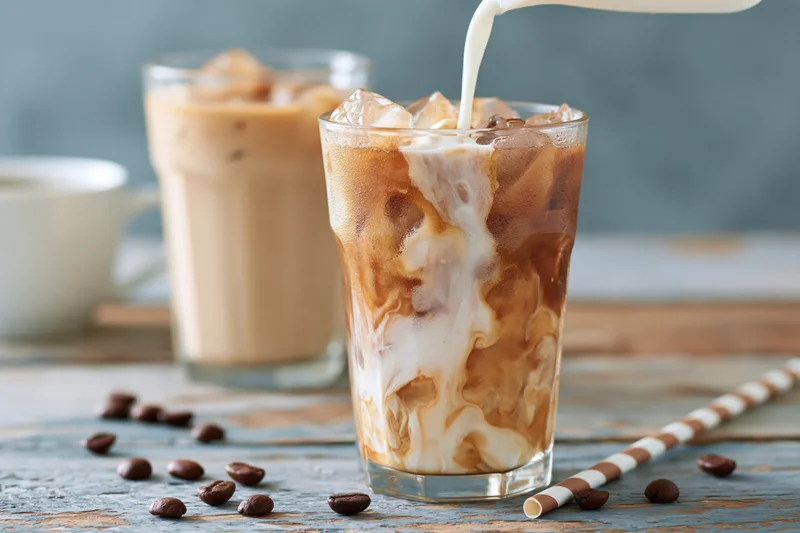 Milk being poured into a tall glass of iced coffee with ice cubes and coffee beans scattered on a wooden table