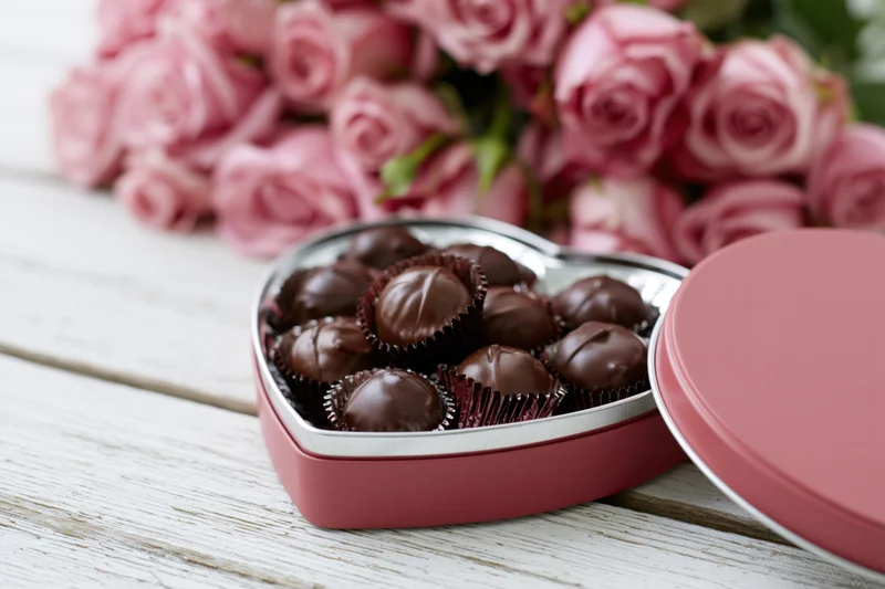 Open heart-shaped box of dark chocolate truffles on wooden table with pink roses in background