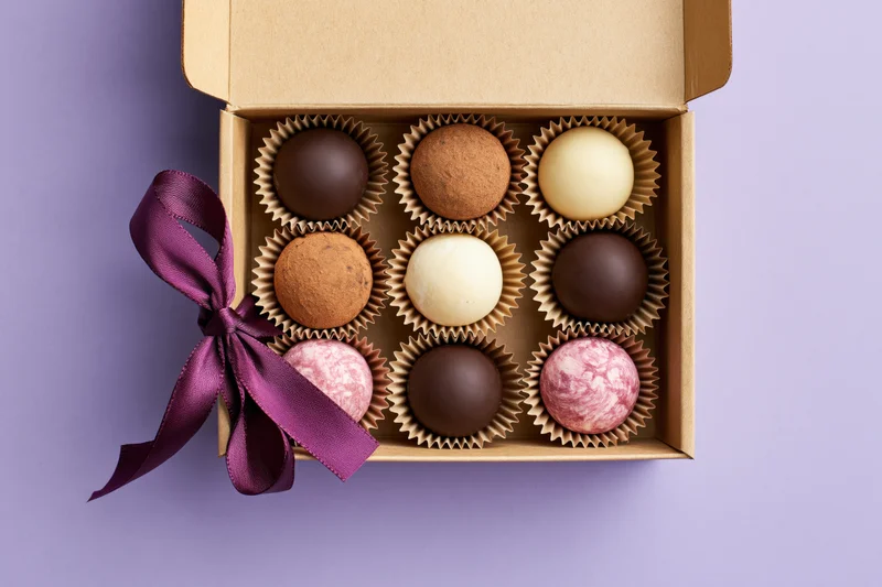 Open kraft cardboard gift box holding nine assorted chocolate truffles arranged in grid with purple ribbon on purple background