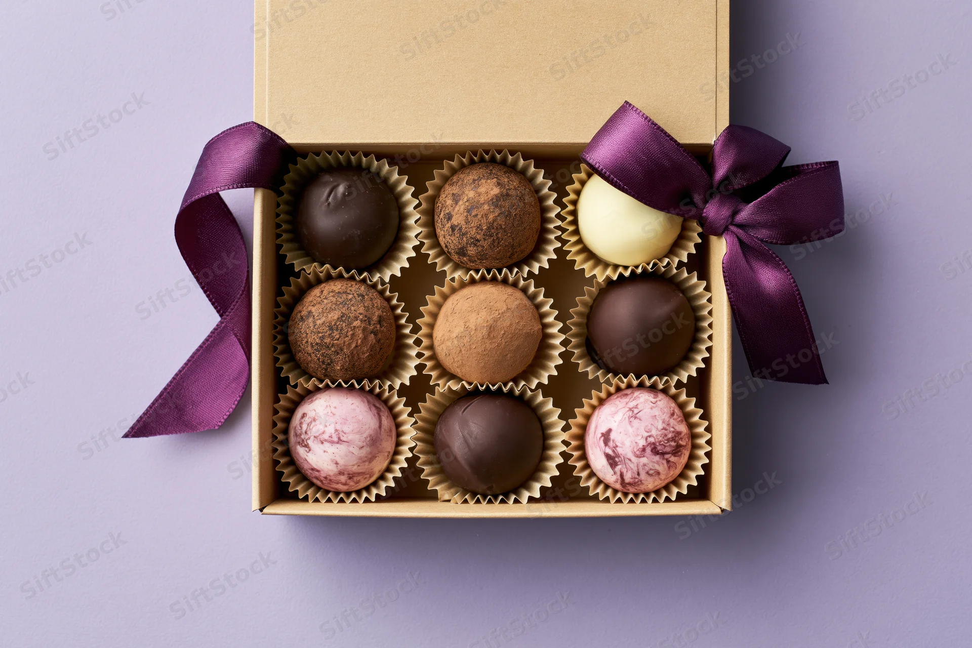 Open kraft gift box with nine assorted chocolate truffles arranged in rows, tied with purple ribbon on purple background