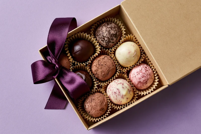 Open kraft cardboard gift box holding nine assorted chocolate truffles arranged in paper liners with a purple ribbon bow on a purple background