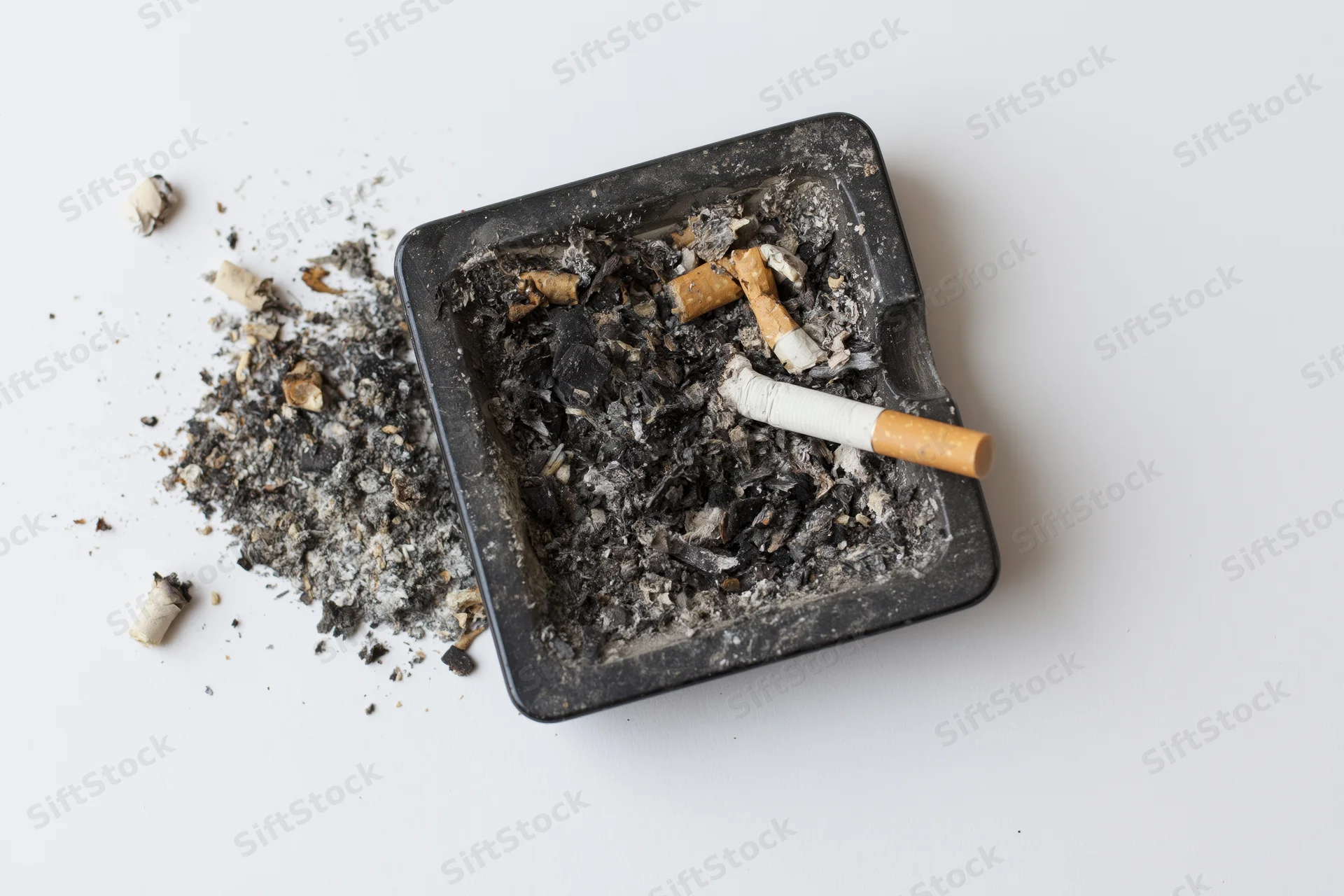 Dirty black square glass ashtray filled with cigarette ash and butts on white surface