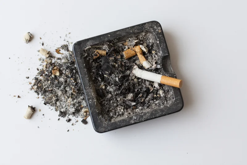 Dirty black square glass ashtray filled with cigarette ash and butts on white surface