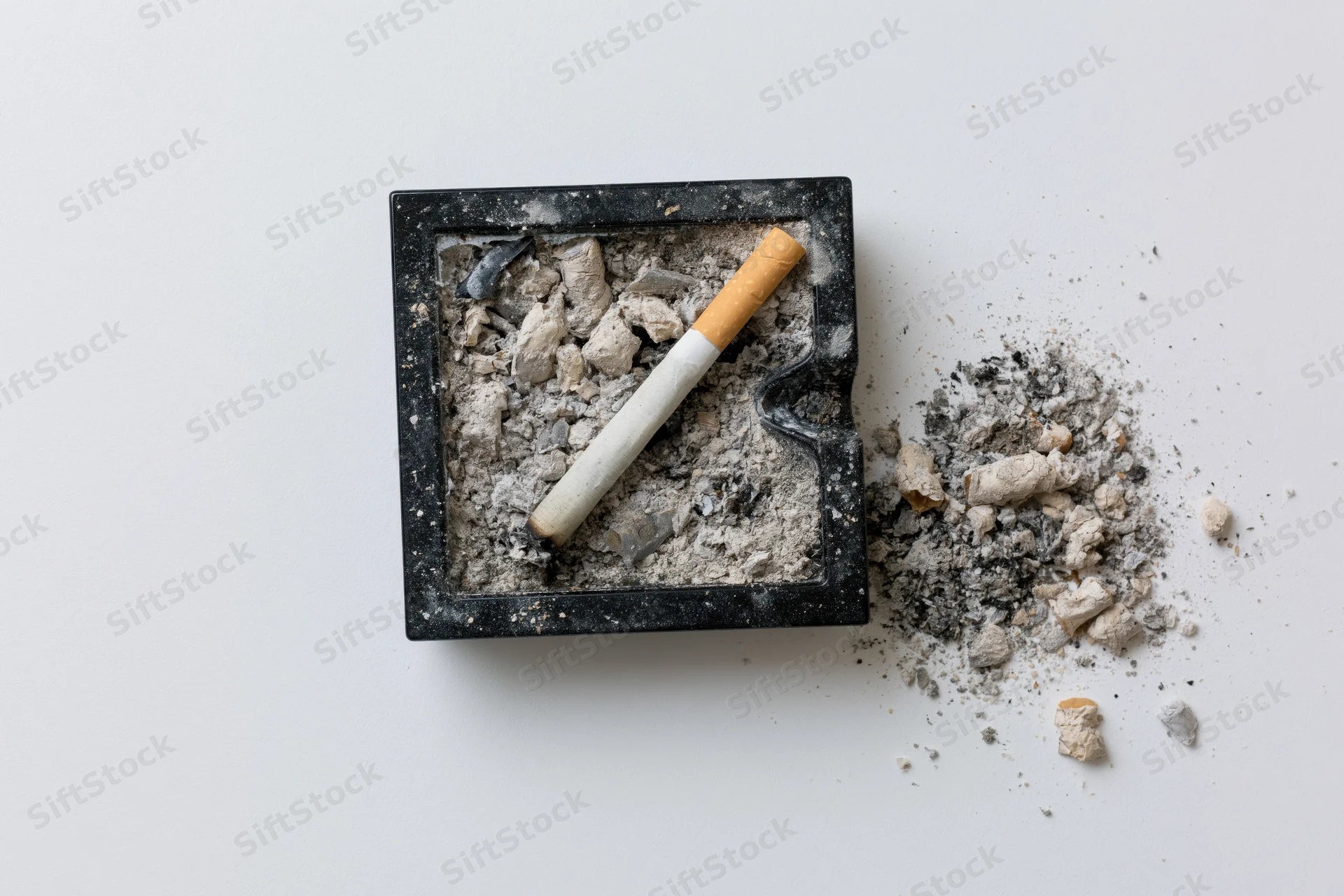 Dirty black square glass ashtray filled with cigarette ash and a butt, viewed from above on white surface