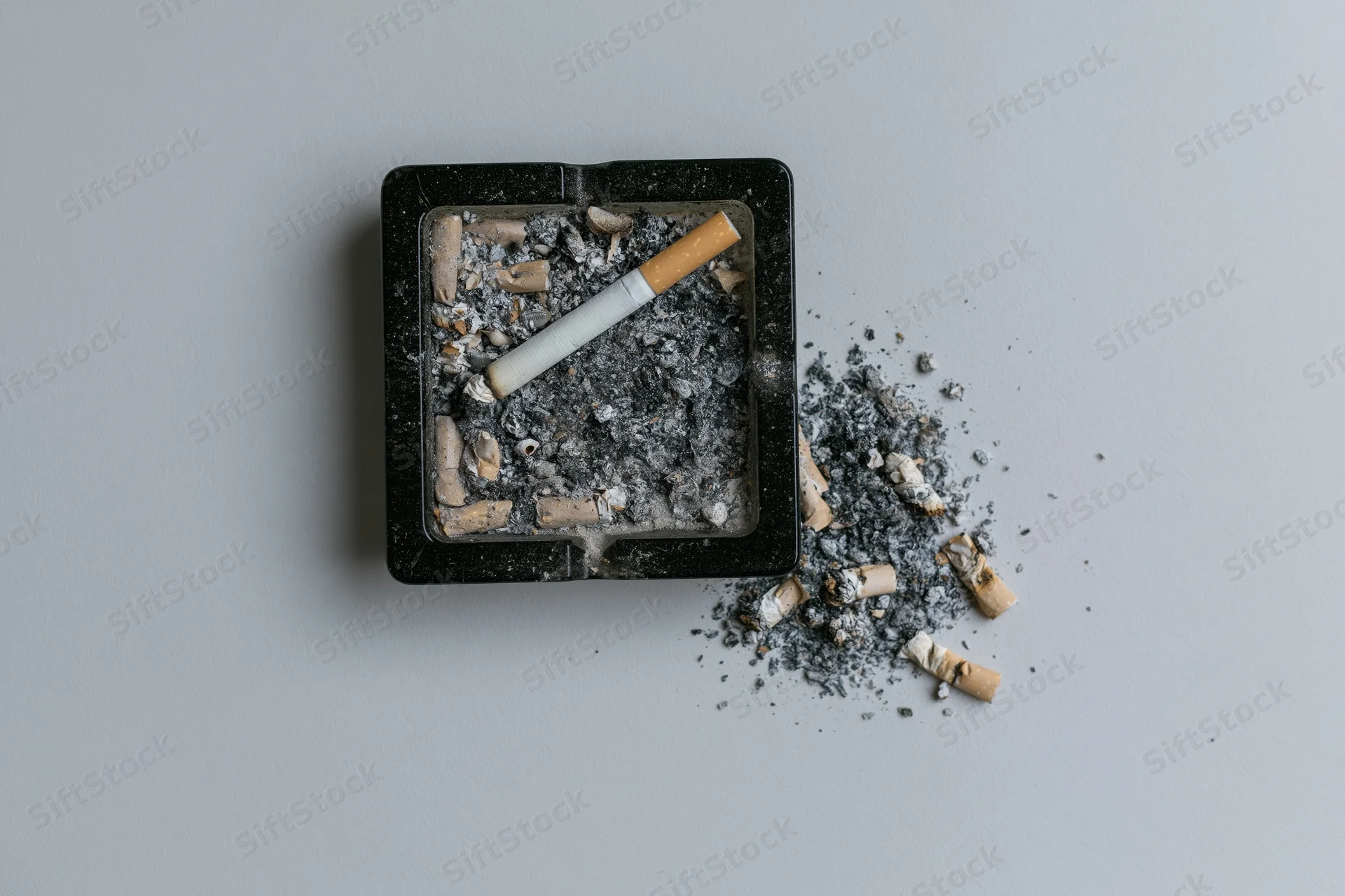 Dirty black square glass ashtray filled with cigarette butts and ash on a plain surface