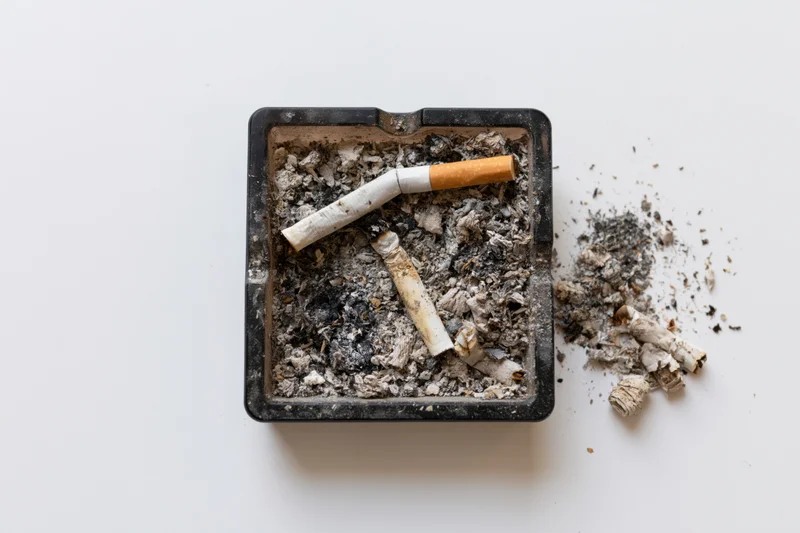 Dirty black square glass ashtray filled with cigarette ash and butts on white background